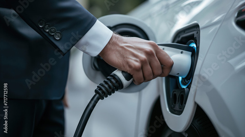 Young business man in suit traveling by electric car at summer, having stop at charging station standing plugging cable