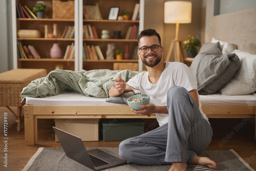 Young man using laptop while having breakfast in bedroom Young man using laptop while having breakfast in bedroom