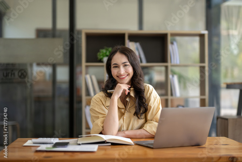 Attractive Asian businesswoman sitting with laptop and documents at table in office, thinking an idea, looking at camera