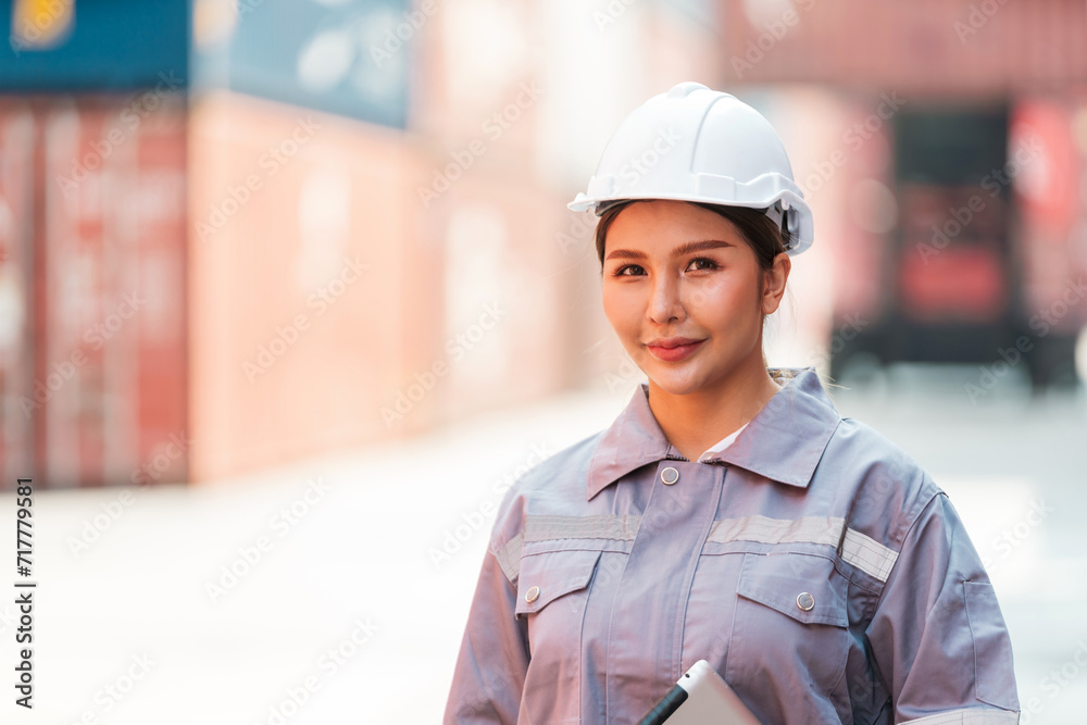 Attractive Thai woman industrial engineer in white hard hat, safety vest with a blurred container yard in background. Working in the logistics center. Inspector, supervisor in container terminal.