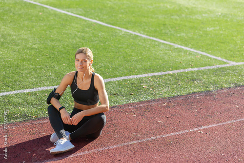 Sporty woman enjoying rest time sitting on track field in sunshine after exercising, showcasing healthy lifestyle.