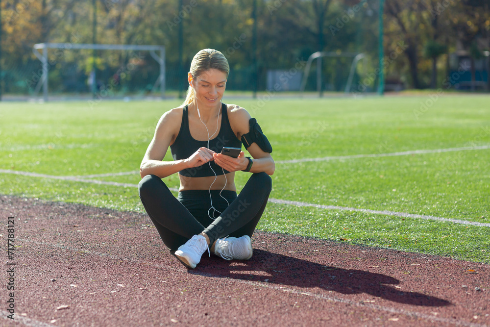 Obraz premium Active young woman enjoys a break on a sports track, choosing music on her phone with earphones on a bright day.