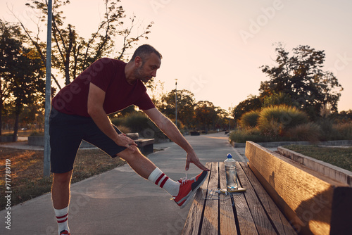 Sporty adult man stretching on a park bench after jogging in sunset time. photo