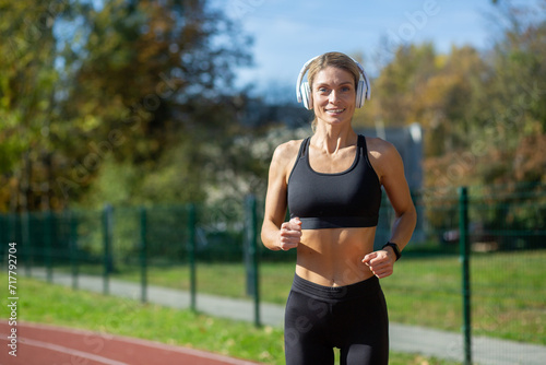 A smiling fit woman with headphones enjoys a run in a sunny park. She exemplifies healthy living and fitness routine.