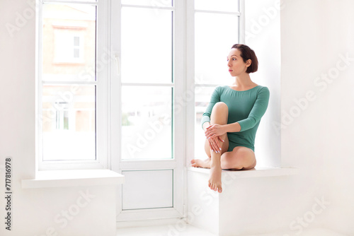 Portrait of beautiful lonely pensive young woman in green body sitting next to large window