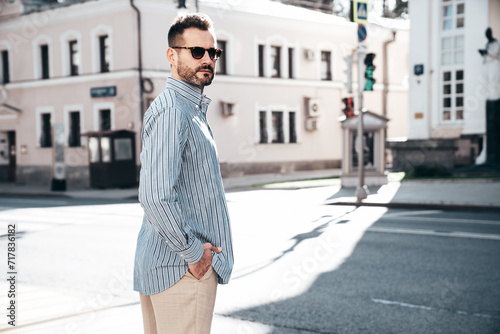 Portrait of handsome confident stylish hipster lambersexual model. Sexy modern man dressed in blue shirt and trousers. Fashion male posing on street background in Europe city at sunset. In sunglasses