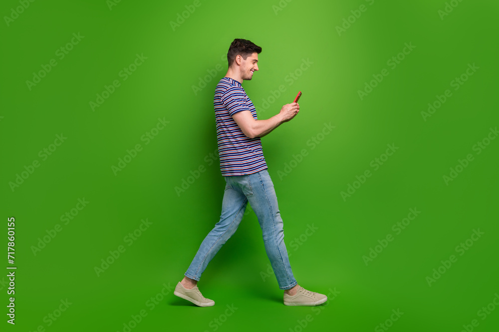 Full length photo of intelligent man wear t-shirt denim pants look at smartphone go to empty space isolated on green color background Full length photo of intelligent man wear t-shirt denim pants look at smartphone go to empty space isolated on green color background