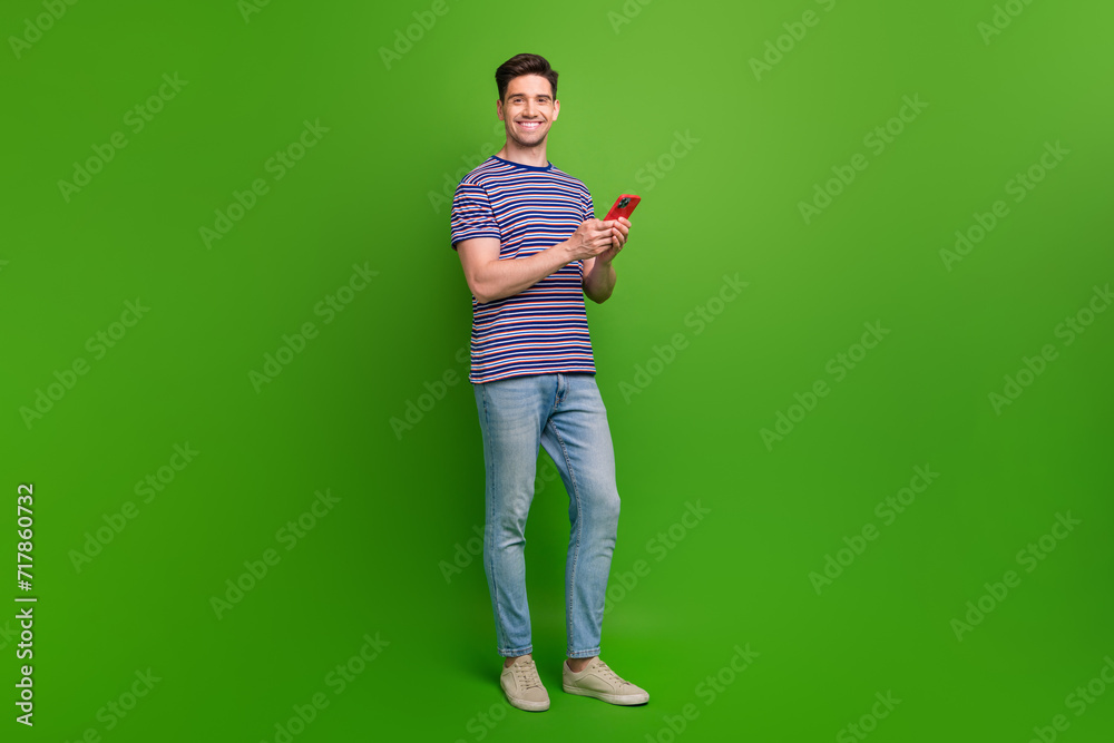 Full length photo of optimistic smart man wear t-shirt denim pants holding smartphone read email isolated on green color background Full length photo of optimistic smart man wear t-shirt denim pants holding smartphone read email isolated on green color background