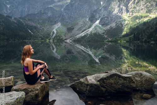 Young blonde fit and sporty woman in sportswear sitting on the rock on the shore and admiring beautiful view of green hills and mountains on Morskie Oko lake, High Tatras, Zakopane, Poland.
