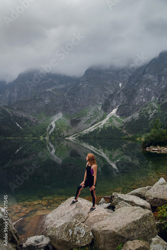 Young beautiful blonde woman in sportswear standing on the rocky shore and enjoying beautiful view of green mountains on Morskie Oko lake, High Tatras, Zakopane, Poland.