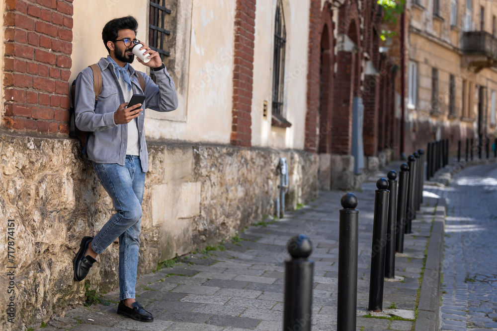 Young handsome hindu man standing near the wall with a coffee cup in hand