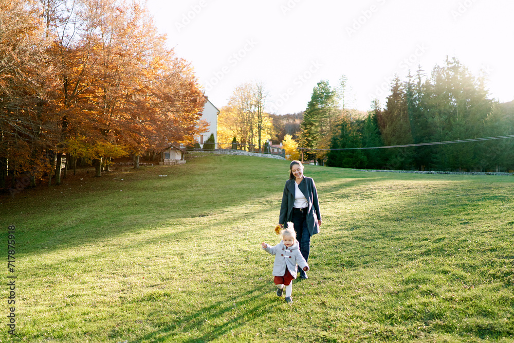 Little girl runs through a green meadow in front of her walking mother
