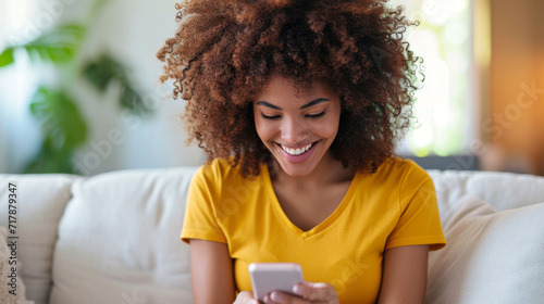 young woman with curly hair, wearing a yellow shirt, smiling and looking down at her smartphone