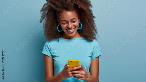 smiling young woman looking down at her yellow smartphone with pleasure, wearing a blue top and matching blue hoop earrings against a soft blue background photo