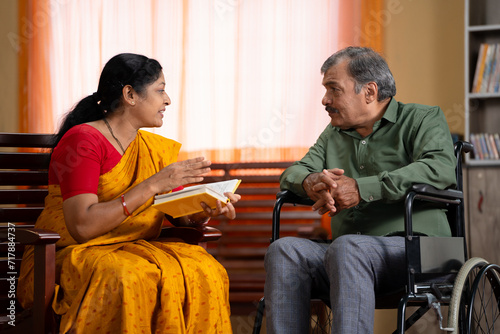 caring Indian senior wife telling by reading novel or story from book to husband sitting on wheelchair at home - concept of family support, relationship bonding and compassion