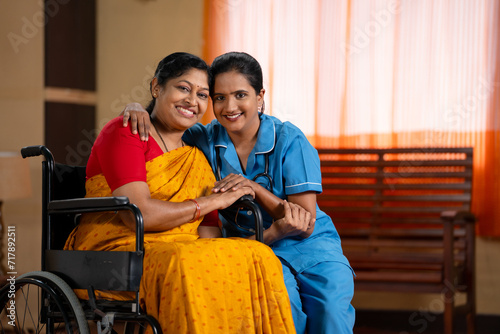 Happy recovered woman on wheelchair with caretaker looking camera at home - concept of professional occupation, healthcare assistance and compassion