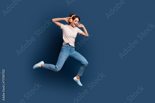Full body photo of pretty young girl running jump excited dance headphones wear trendy white outfit isolated on dark blue color background © deagreez