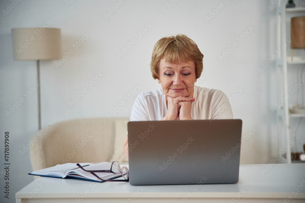 Front view. Sitting by table with laptop. Senior elderly woman is at home in the living room Front view. Sitting by table with laptop. Senior elderly woman is at home in the living room