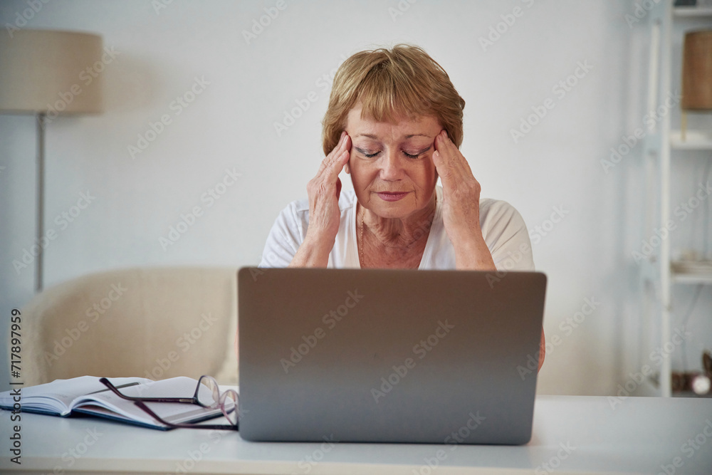 Headache, sitting by laptop. Senior elderly woman is at home in the living room Headache, sitting by laptop. Senior elderly woman is at home in the living room