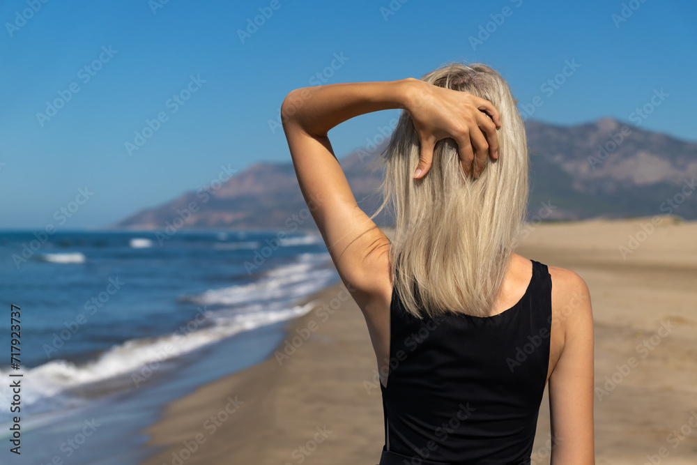 Slender blonde young woman in a black one-piece swimsuit on a sandy beach holding her hair with her hand, rear view. Concept of freedom, travel, lifestyle