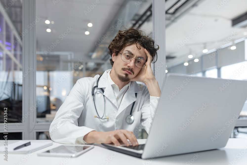 A man in a white medical coat with a stethoscope sits in front of a laptop computer in a medical office. Doctor thinking using computer A man in a white medical coat with a stethoscope sits in front of a laptop computer in a medical office. Doctor thinking using computer