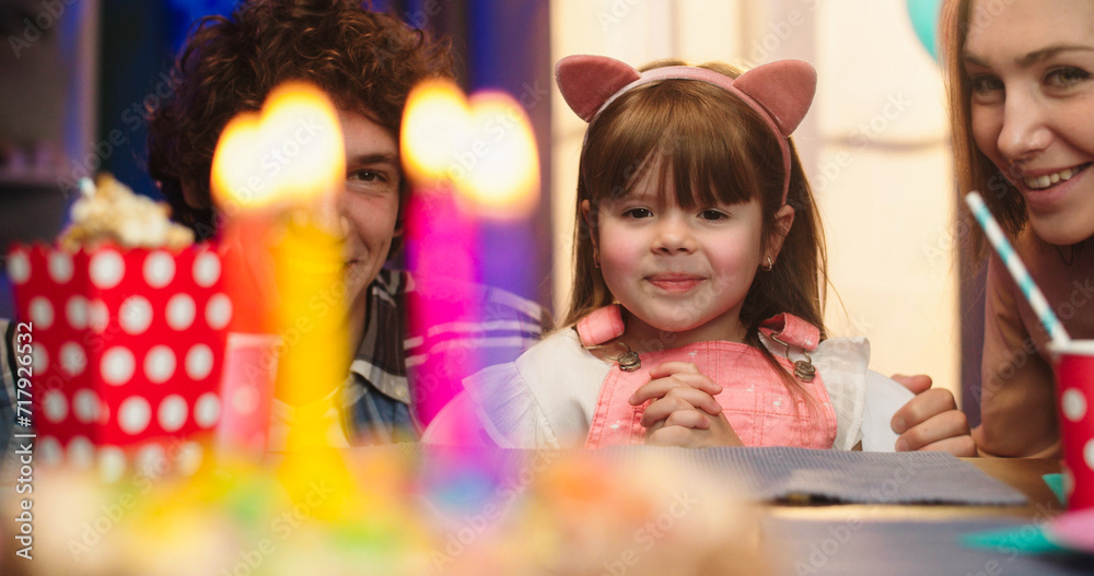 Portrait shot of the cute small Caucasian girl sitting in front of the birthday cake with lighted candles between her parents and wishing something before blow candles.