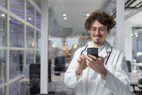 A man in a white lab coat and stethoscope holds a cell phone inside a medical office of a clinic. photo
