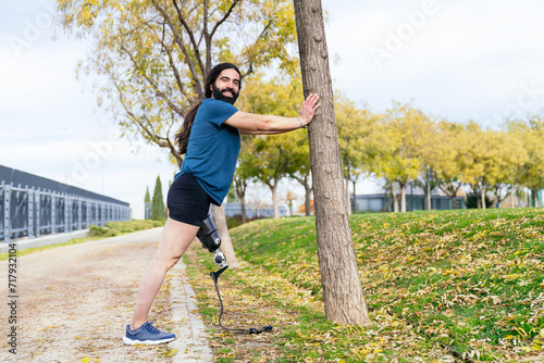 Amputee athlete prepares for a jog, stretching his prosthetic leg against a tree in a leaf-strewn park.