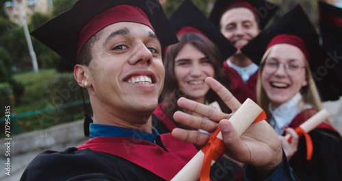 Fotografie Close up of the mulatto young joyful guy in academic cap and gowns taking funny selfie photos with his funny and happy friends students at their graduation day