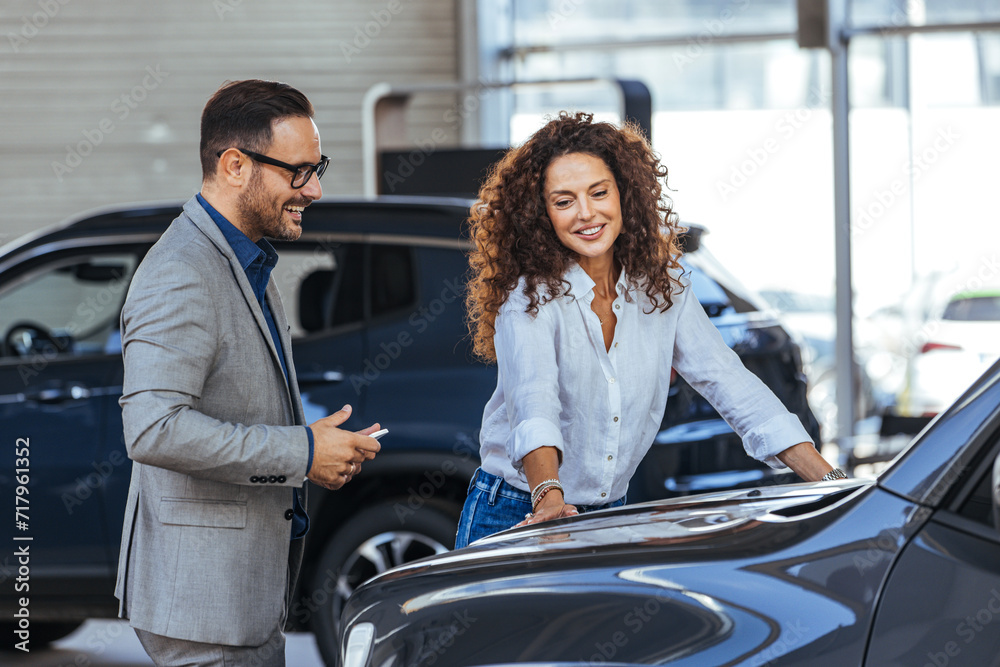 Happy woman with car dealer in auto show or salon. Car Sales Manager Showing Auto To Lady Buyer. Beautiful caucasian female client customer choosing new car.