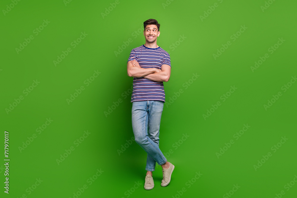 Full length photo of positive man wear t-shirt denim pants arms folded look at promo empty space isolated on green color background Full length photo of positive man wear t-shirt denim pants arms folded look at promo empty space isolated on green color background