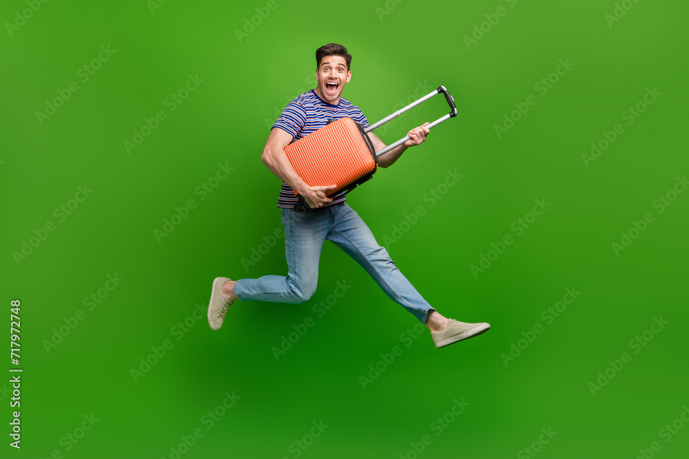 Full length photo of ecstatic man wear stylish t-shirt jeans trousers jumping with baggage in hands isolated on green color background Full length photo of ecstatic man wear stylish t-shirt jeans trousers jumping with baggage in hands isolated on green color background