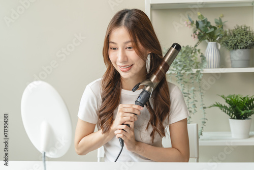 women hairdo makeup routine, Young woman looking at the mirror and using iron hair curling curly long hair at home. photo