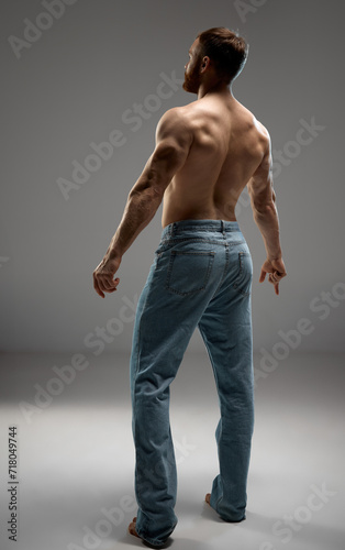 Rear view portrait of strong male model with healthy back posing in denim jeans against grey studio background. Men's skincare promotion focusing on natural body care. Concept of beauty and fashion.