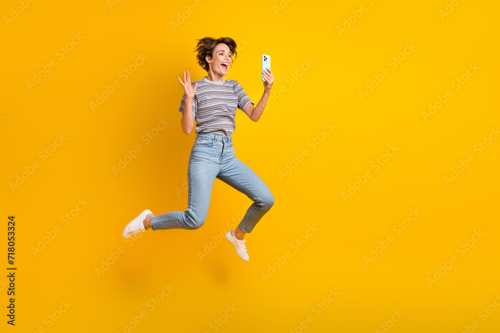 Full body photo of overjoyed energetic lady jump hold smart phone make selfie waving hi empty space isolated on yellow color background