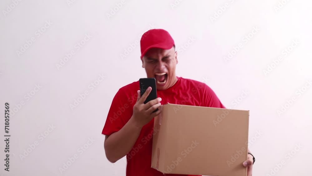 Surprised young asian courier man holding cardboard while received good news on his phone