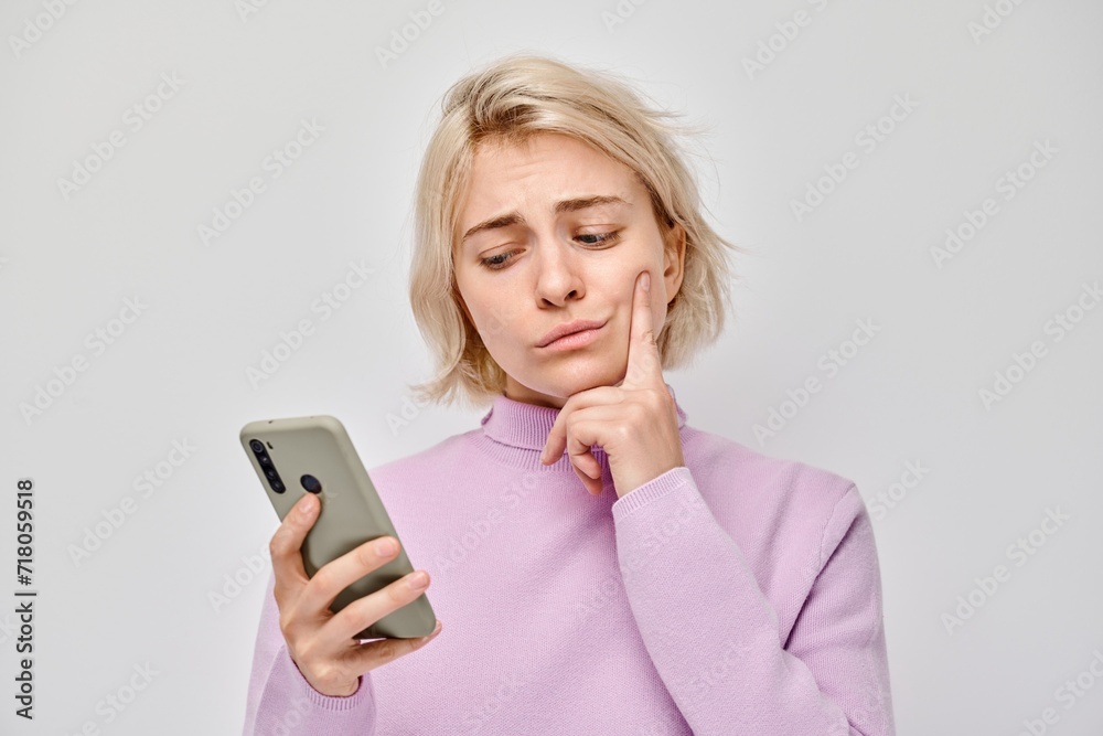 Portrait of young blond woman looks on mobile phone and thinks. Person with smartphone isolated on white background. Portrait of young blond woman looks on mobile phone and thinks. Person with smartphone isolated on white background.
