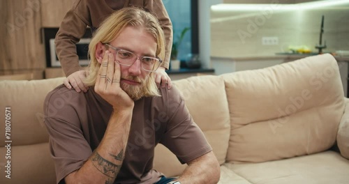 A tortured blond man with a beard and glasses sits on the sofa while his little albino son pesters him and wants to play with the man at home in a modern apartment. Tired man with glasses cannot pay photo
