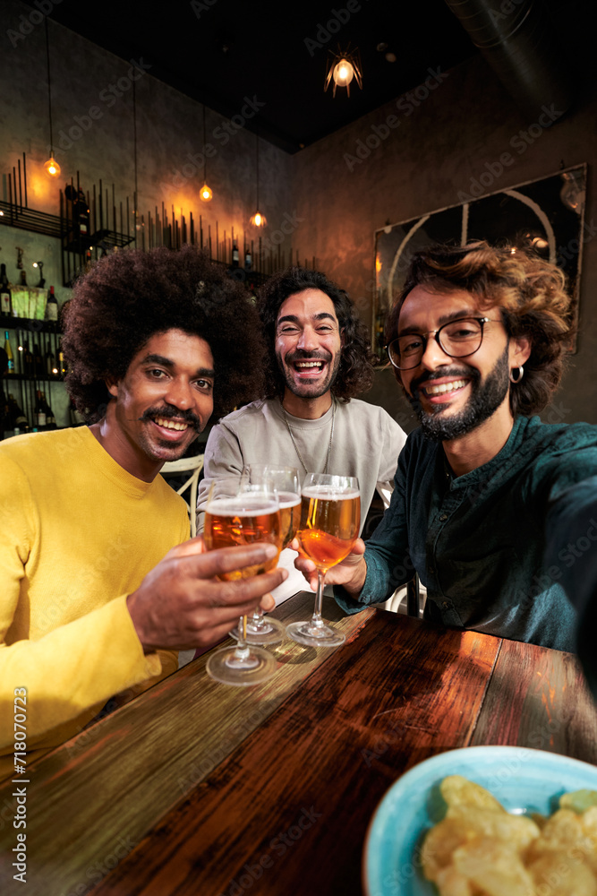 Cheerful vertical photo of three men multiethnic friends toasting with beers in a bar, looking at camera smiling while taking a selfie to post on social media apps. Copy space.