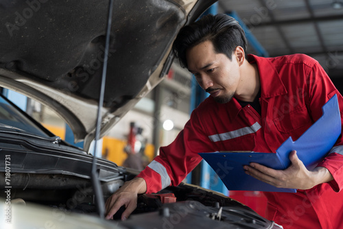 Male mechanic holding clipboard during working and checking car engine under hood of car in auto car repair service garage. Male technician inspecting car engine in garage