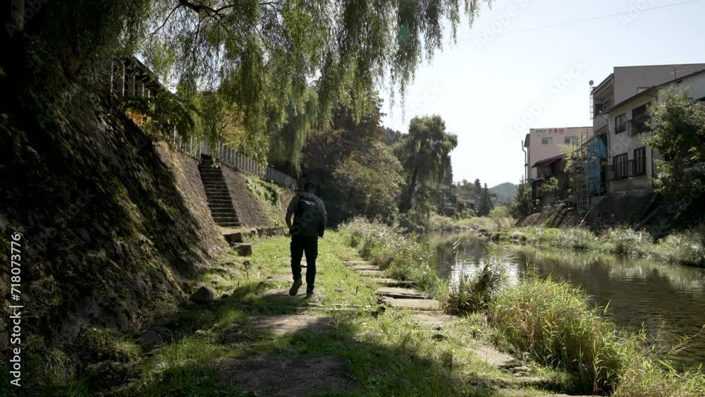 Solo Male Backpacker Walking Away along the path beside the Miyagawa River in Takayama. Static Shot Solo Male Backpacker Walking Away along the path beside the Miyagawa River in Takayama. Static Shot