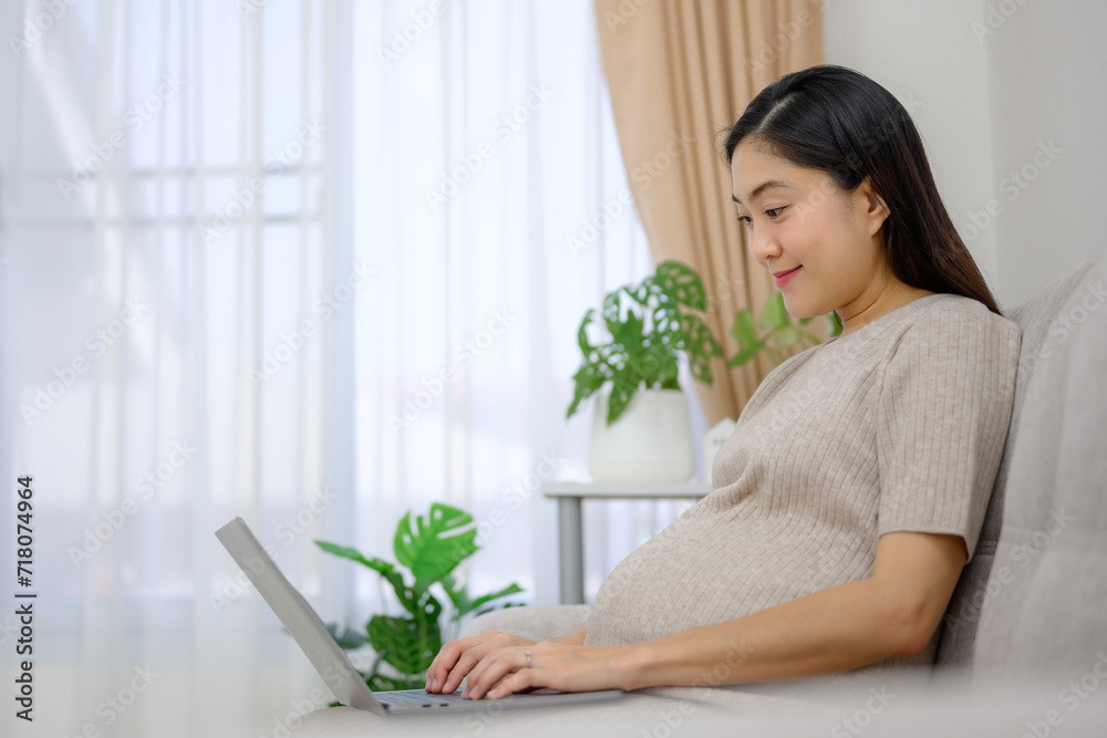 Asian pregnant woman using laptop doing freelance work on sofa in living room at home Carrying a large belly Happy pregnant woman at her home before giving birth Asian pregnant woman using laptop doing freelance work on sofa in living room at home Carrying a large belly Happy pregnant woman at her home before giving birth