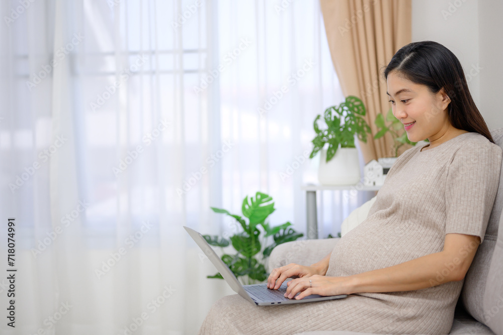 Asian pregnant woman using laptop doing freelance work on sofa in living room at home Carrying a large belly Happy pregnant woman at her home before giving birth Asian pregnant woman using laptop doing freelance work on sofa in living room at home Carrying a large belly Happy pregnant woman at her home before giving birth