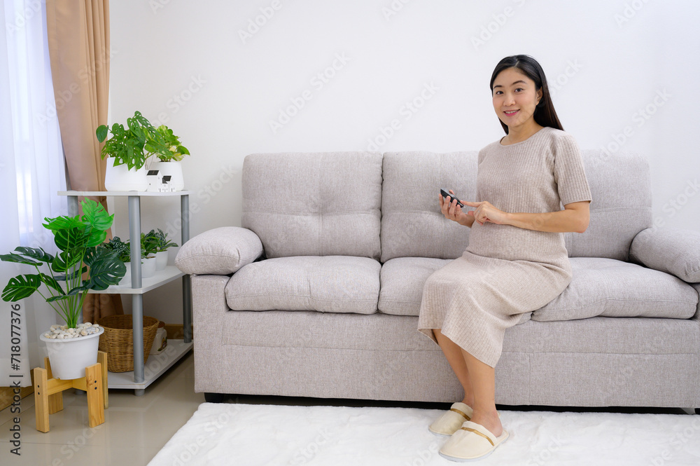 Asian pregnant woman with diabetes A pregnant woman sits checking her blood sugar level on the sofa in the living room at home