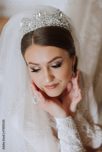 Beautiful, young, perfect model bride in a white lace dress sits in the room, bedroom in the morning, putting an earring on her ear. Wedding photography, portrait, concept.