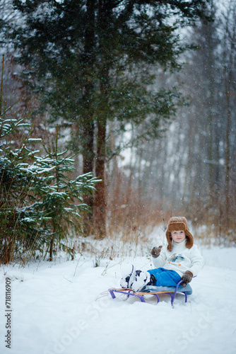 A 4-year-old boy sits on a sled in a snowy forest in winter. A warm brown hat, blue pants, a white fur collar