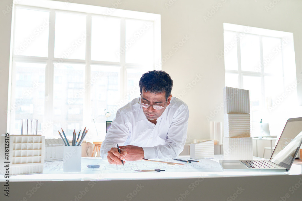 Portrait of male Indian architect drawing plans at workstation in white office interior, copy space