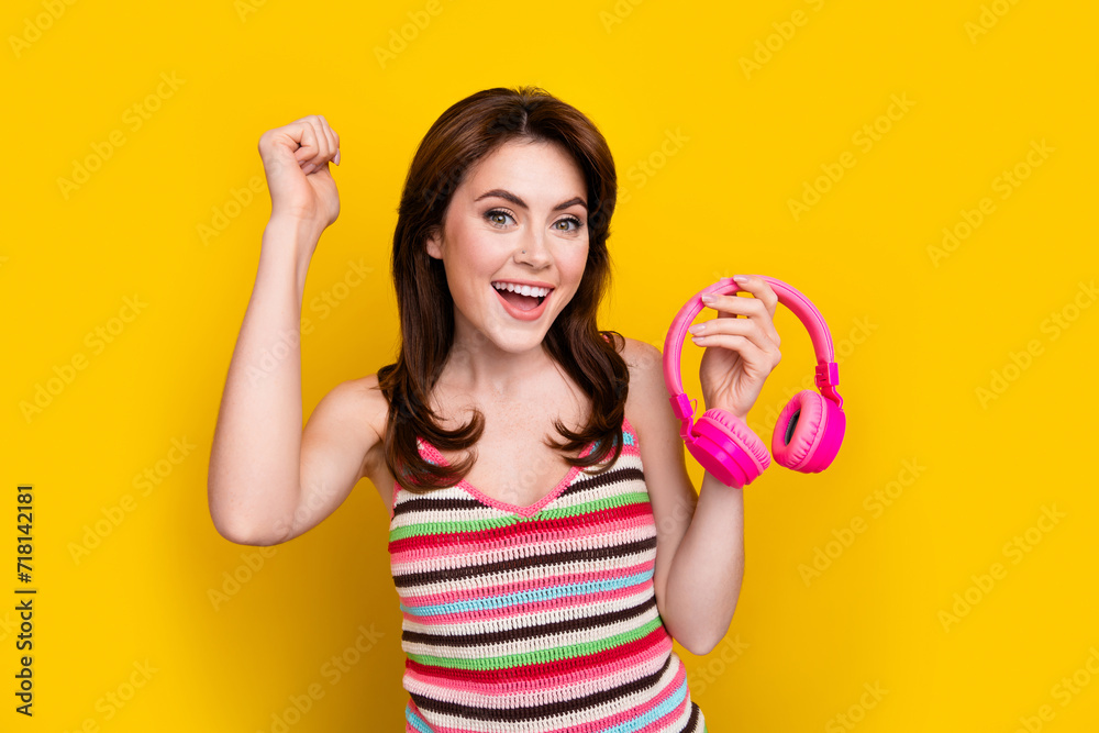 Photo of satisfied good mood cute girl dressed striped top hold headphones clenching fist isolated on bright yellow color background Photo of satisfied good mood cute girl dressed striped top hold headphones clenching fist isolated on bright yellow color background