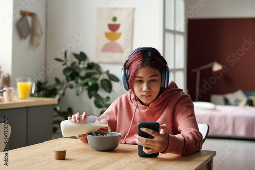 Bild auf Leinwand Cute Asian female teenager in headphones pouring fresh milk into bowl with cornf