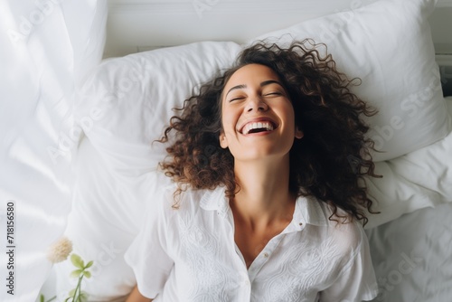 A woman smiles brightly, lying in a bed, exuding happiness and comfort in the morning light.